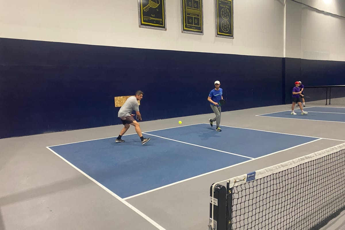 Three players mid-rally on the courts at Dinkers, the ball in the air over the net
